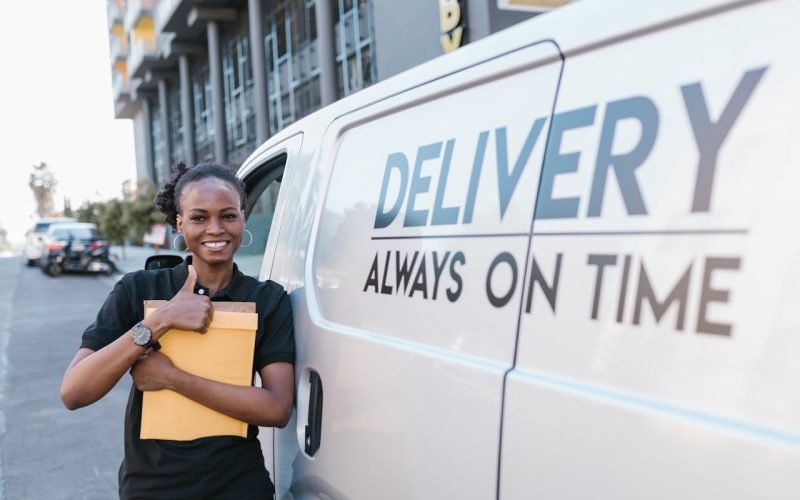 A cheerful delivery woman standing by a van with a parcel, ready for delivery.